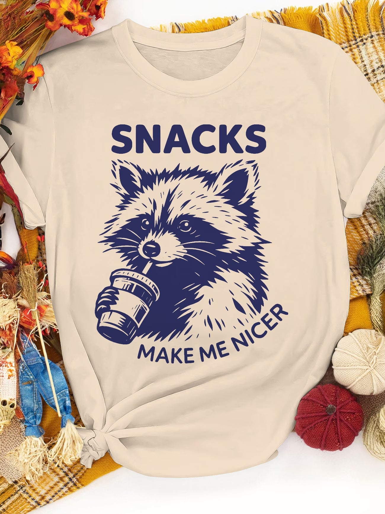 Beige t-shirt with a raccoon graphic and text 'SNACKS MAKE ME NICER' on a decorative background.