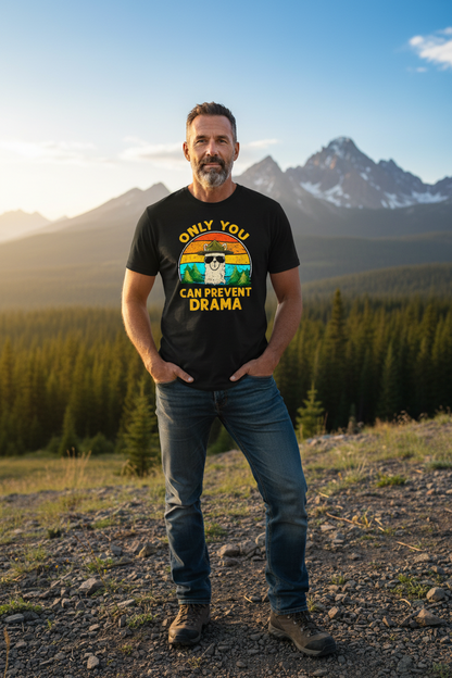 Man wearing a t-shirt with a mountain and text design, standing in a scenic outdoor setting with mountains and trees.