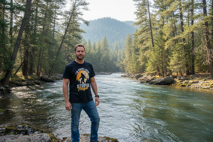 Man standing by a river with trees and mountains in the background