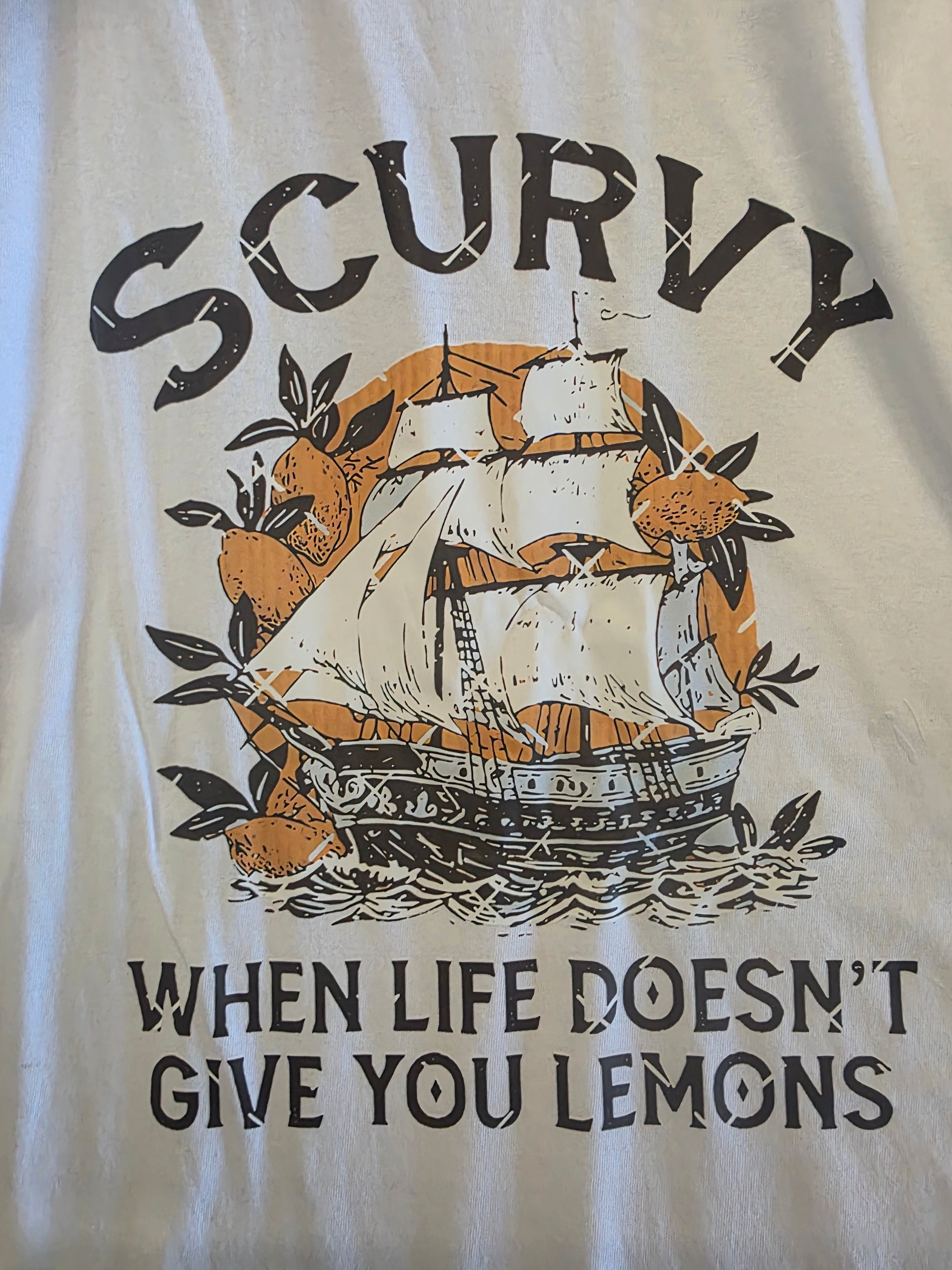 A close-up of a white graphic t-shirt featuring a vintage-style print of a sailing ship, lemons, and waves. The text reads 'SCURVY WHEN LIFE DOESN'T GIVE YOU LEMONS' in black lettering.