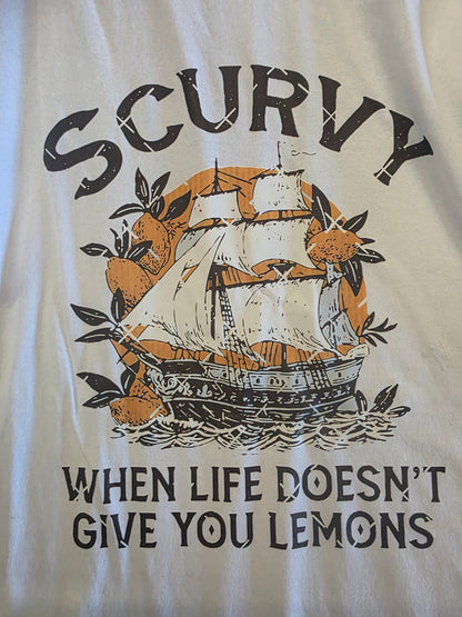 A close-up of a white graphic t-shirt featuring a vintage-style print of a sailing ship, lemons, and waves. The text reads 'SCURVY WHEN LIFE DOESN'T GIVE YOU LEMONS' in black lettering.