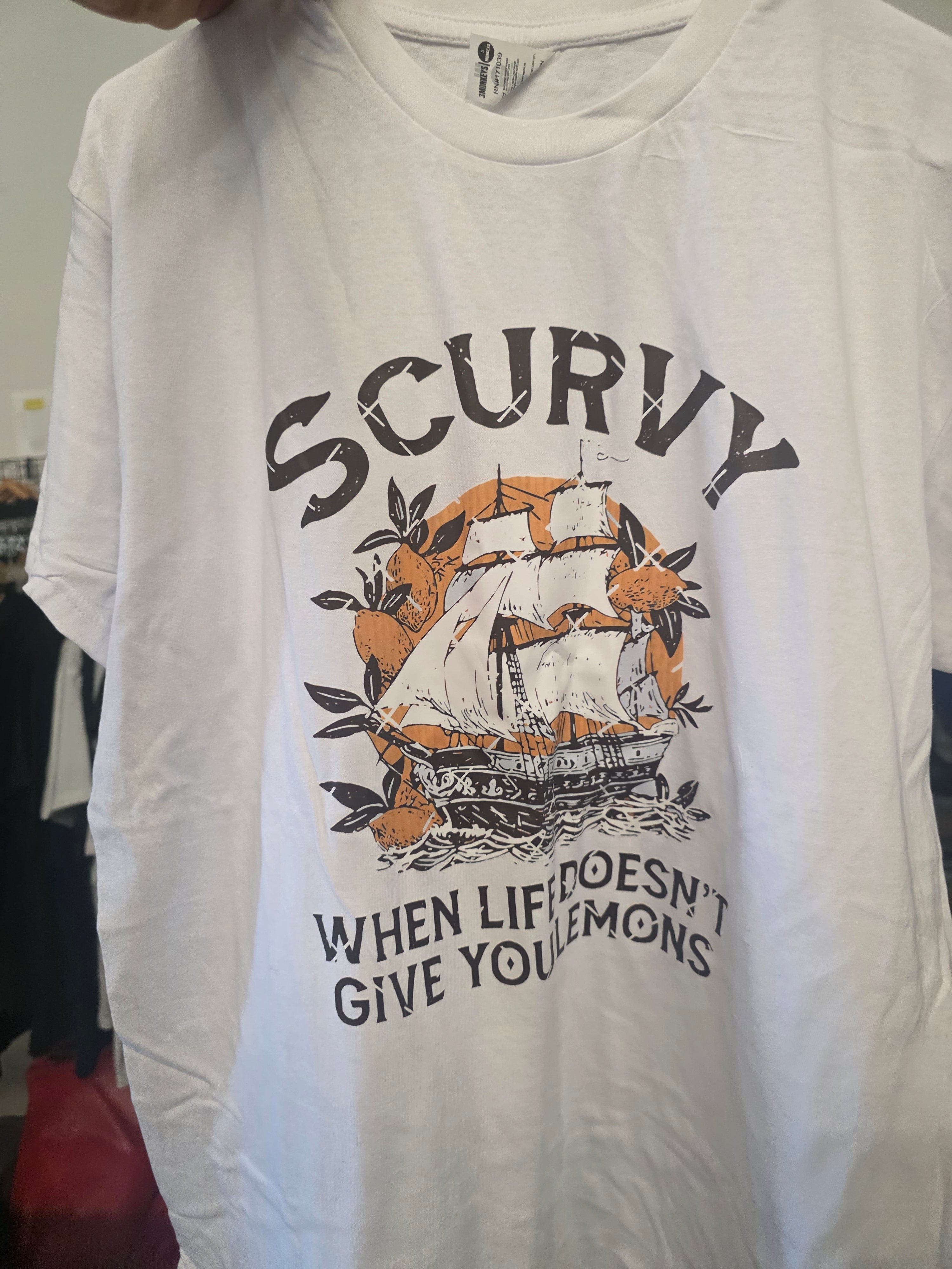 A white short-sleeve graphic t-shirt featuring a vintage-style print of a sailing ship surrounded by oranges and the text 'SCURVY WHEN LIFE DOESN'T GIVE YOU Lemons'.